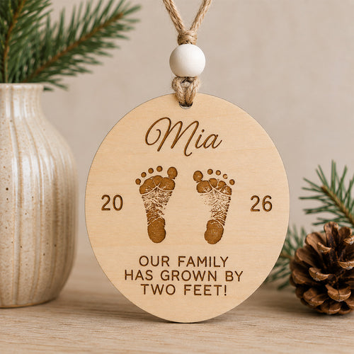 Personalized baby's first Christmas ornament featuring engraved baby footprints, a custom name, birth year, and the message "Our family has grown by two feet!" handcrafted from Baltic birch.